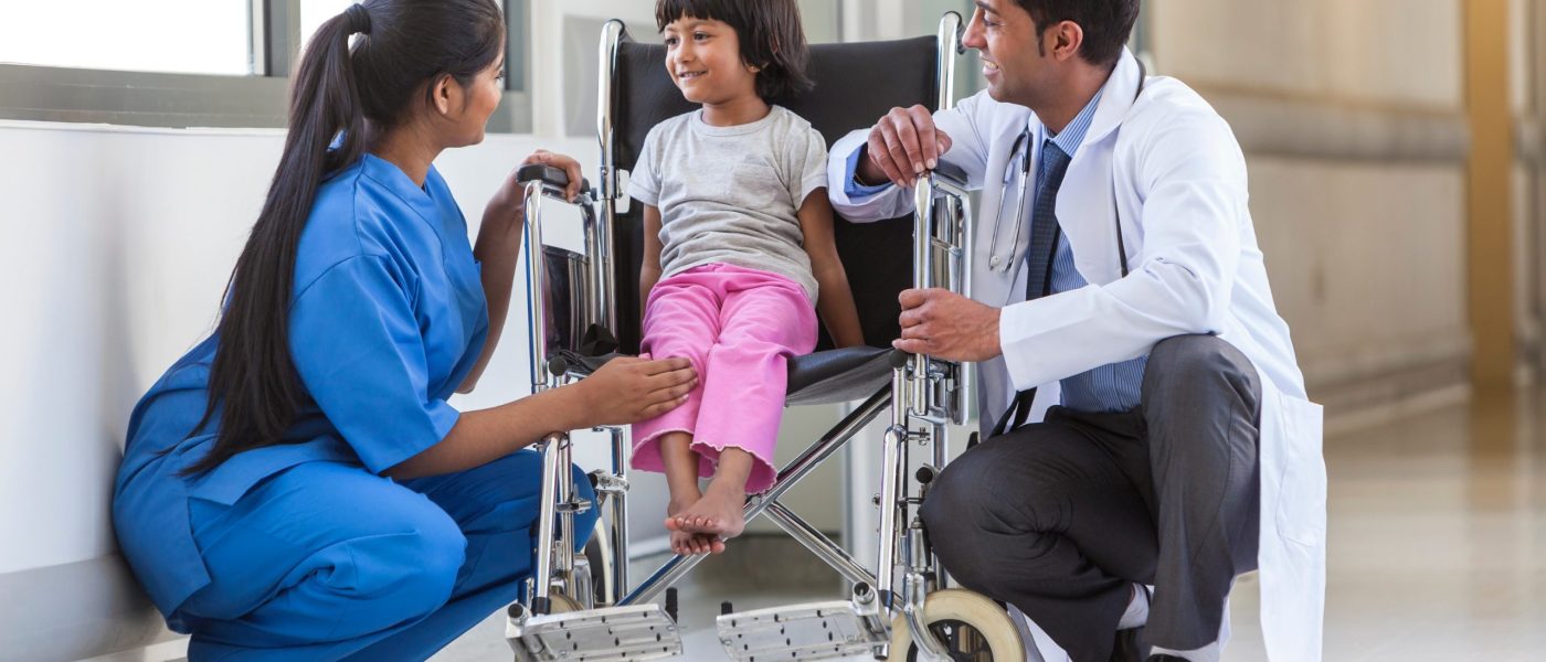 Young female child patient in wheelchair sitting in hospital corridor with Indian Asian female nurse and male doctor